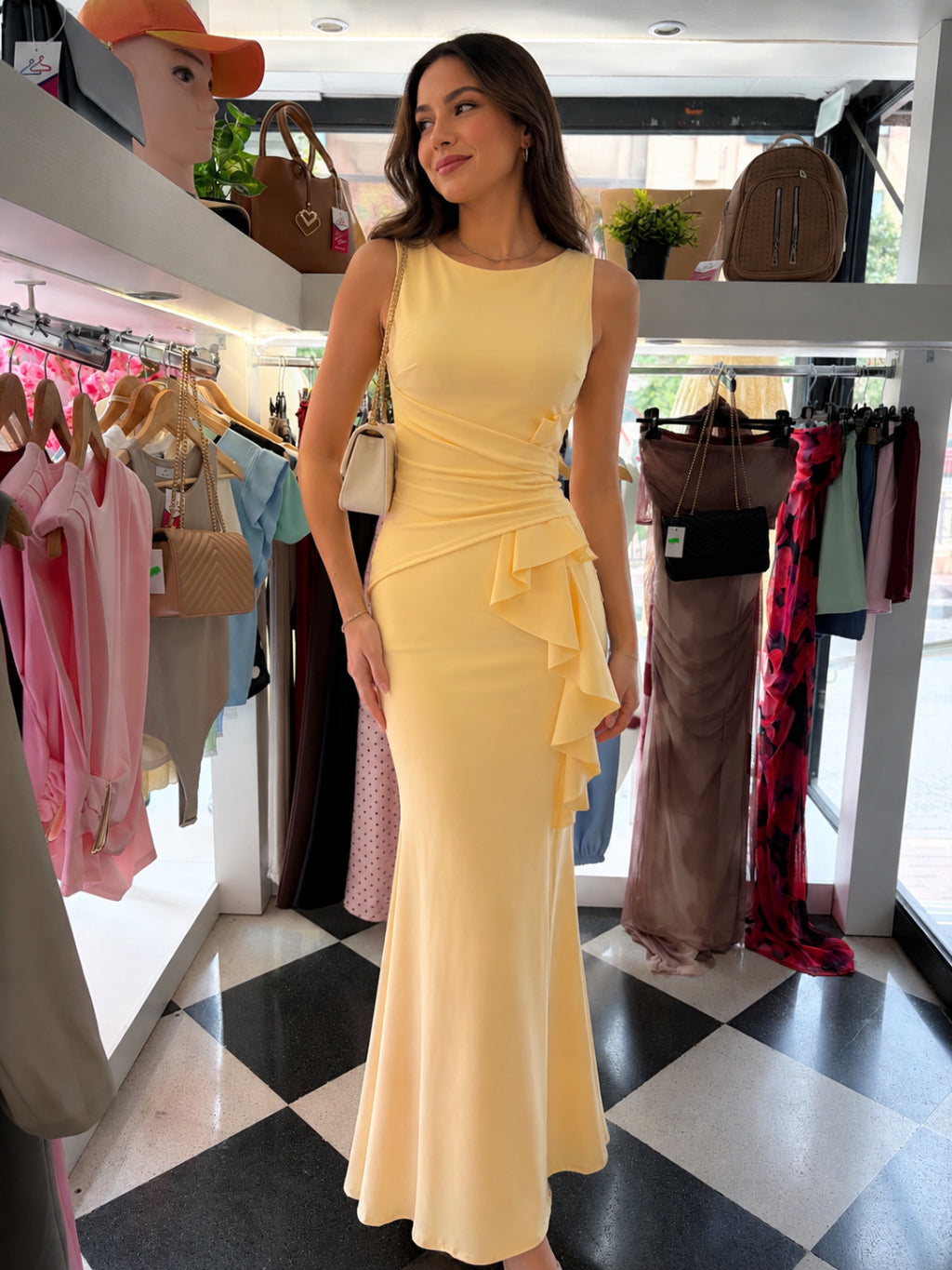 Woman in a yellow dress standing in a clothing store with racks of clothes and bags in the background.
