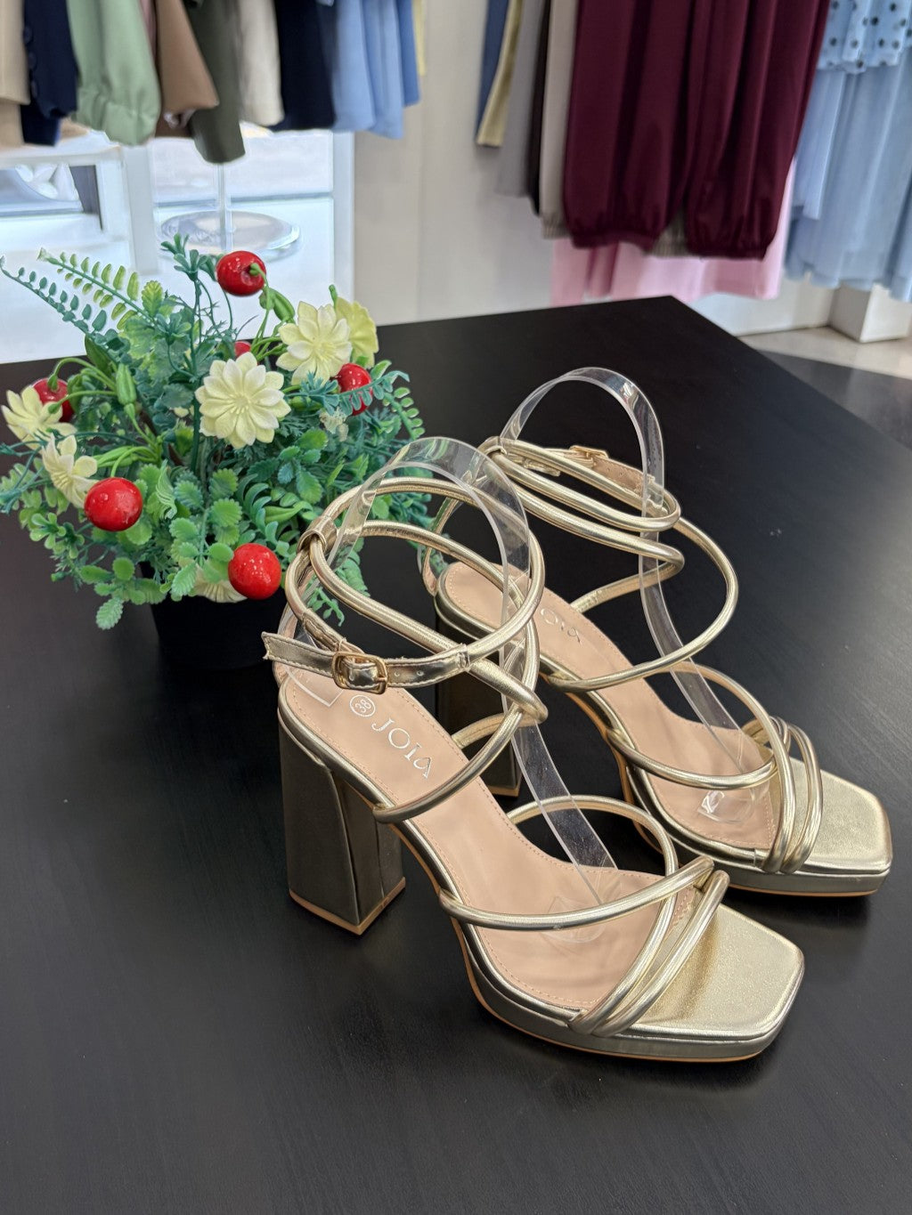 Gold high-heeled sandals on a dark surface with a small potted plant in the background.