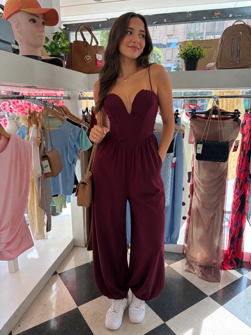 Woman in a maroon jumpsuit standing in a clothing store with racks of clothes and bags in the background.